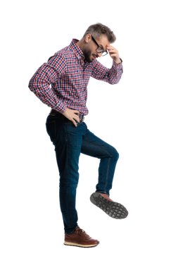 Beyaz arka planda ayakkabı tabanını incelerken ekose gömlekli, alınlı, rahatsız bir adamın yan görünüşü.