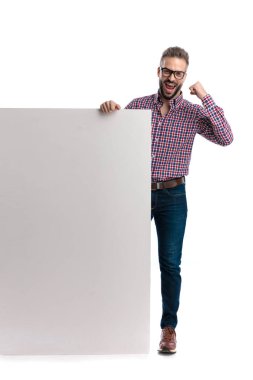 Gözlüklü ve ekose gömlekli heyecanlı bir adam yumruğunu kaldırdı ve başarısını büyük boş bir reklam panosuyla kutlarken beyaz arka planda ileriye bakıyordu.