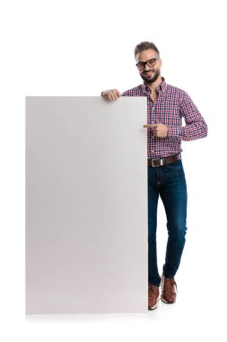 Gözlüklü ve ekose gömlekli neşeli bir adam büyük boş bir reklam panosuna yaslanıyor ve beyaz arka planda ileriye bakarken işaret ediyor.