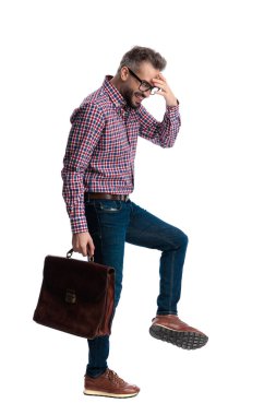 Utanmış sakallı bir adamın evrak çantasını tutarken yürümesi ve beyaz arka planda izole edilmiş alnına dokunması.