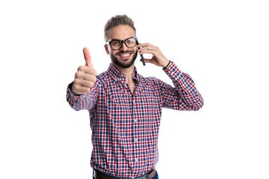 Ekose gömlekli ve gözlüklü mutlu bir adam cep telefonuyla konuşurken başparmak hareketi yapıyor ve beyaz arka planda izole edilmiş bir şekilde ileriye bakıyor.