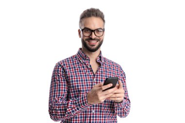 Ekose gömlek ve gözlük takan neşeli genç adam beyaz arka planda mesajlaşırken ve ileriye bakarken akıllı telefon tutuyor.