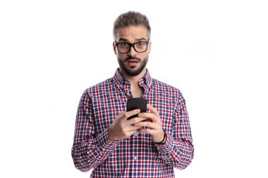 Beyaz arka planda afallamış bir ifadeyle ileriye bakarken iki elinde cep telefonu tutan şaşkın bir adam.