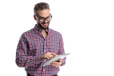 Ekose gömlek ve gözlük takan mutlu adam tablet tutuyor ve beyaz arka planda gülümserken ekrana dokunuyor.