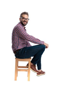 Gözlüklü, ekose gömlekli, tahta bir taburede oturan ve beyaz arka planda izole bir şekilde ileriye bakan mutlu bir adamın yan görüntüsü.