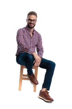 Gözlüklü ve kareli gömlekli mutlu bir adam tahta bir taburede oturuyor gülümsüyor ve beyaz arka planda izole bir şekilde ileriye bakıyor.