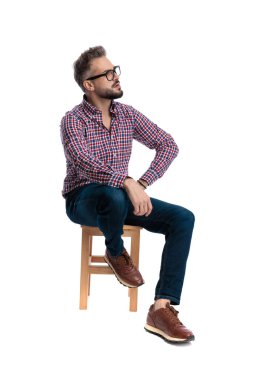 Düşünceli, gözlüklü, kareli tişörtlü, tahta bir taburede oturan ve beyaz arka planda izole bir şekilde başka tarafa bakan bir adamın yan görüntüsü.