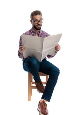Gözlüklü, ekose gömlekli, elinde gazete olan ve beyaz arka planda izole edilmiş tahta bir taburede oturan genç şoka uğramış bir adam.