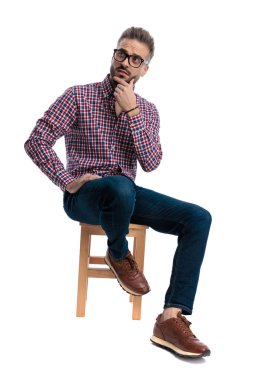 Düşünceli, gözlüklü ve ekose gömlekli genç bir adam tahtadan bir taburede oturuyor ve düşünürken yukarı bakıyor ve beyaz arka planda izole bir şekilde duruyor.
