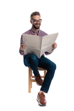 Gözlüklü, ekose gömlekli, elinde gazete tutan, gülümseyen ve beyaz arka planda izole edilmiş tahta bir taburede oturan mutlu bir adam.