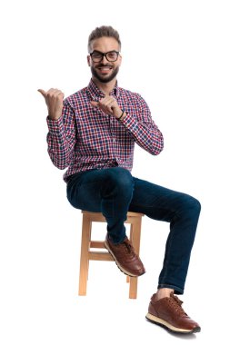 Gözlüklü ve ekose gömlekli neşeli genç bir adam tahta bir taburede oturuyor ve beyaz arka planda izole bir şekilde ileriye bakarken başparmaklarını iki tarafa işaret ediyor.