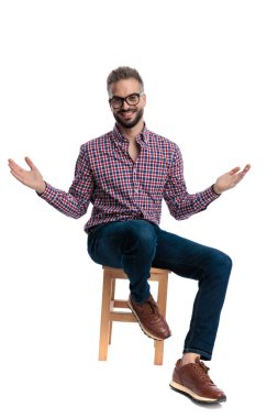 Gözlüklü ve ekose gömlekli neşeli genç adam, tahta bir sandalyede oturuyor ve beyaz arka planda izole edilmiş kameraya gülümserken kollarını açıyor.