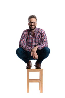 Ekose gömlek ve kot pantolon giyen neşeli genç adam ahşap bir tabureye çömelmiş gülümsüyor ve beyaz arka planda yalnız başına ileriye bakıyor.