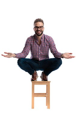 Ekose gömlek ve kot pantolon giyen, kolları geniş bir tahta tabureye çömelmiş, beyaz arka planda ileriye bakan genç adamı selamlıyoruz.