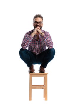 Ahşap bir tabureye çömelmiş, elleri sımsıkı bağlanmış, gülümseyen ve beyaz arka planda ileriye bakan neşeli sıradan bir adam.