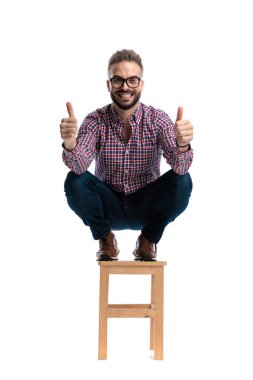 Ahşap bir taburede çömelmiş mutlu bir adam iki eliyle baş parmağını kaldırıyor ve beyaz arka planda ileriye bakarken gülümsüyor.