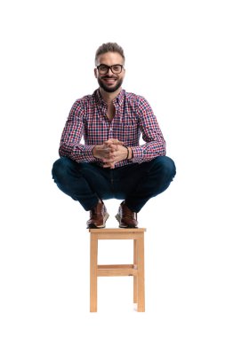 Gözlüklü ve ekose gömlekli neşeli bir genç adam, beyaz arka planda izole edilmiş elleri birlikte, tahta bir taburede çömeliyor.
