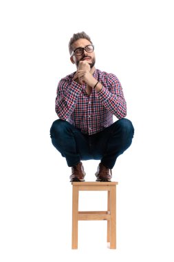 Düşünceli sıradan bir adam, tahta bir tabureye çömelmiş, çenesine dokunuyor ve beyaz arka plana bakarken düşünüyor.