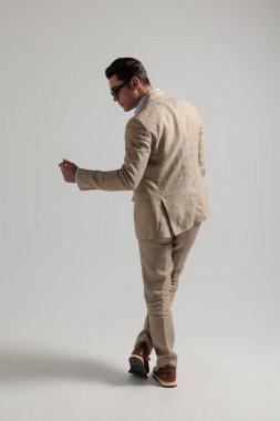 Güneş gözlüğü takan, takım elbiseli, parmaklarını şıklatan ve gri arka plana bakan şık bir adam.