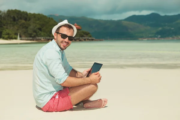 Man sitter på stranden med benen korsade läsning från ipad — Stockfoto