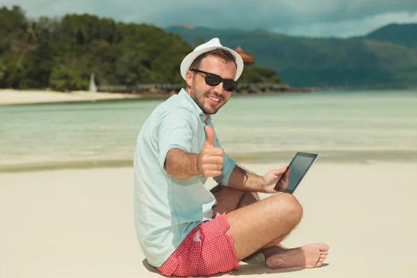 Чоловік дає великі пальці вгору, сидячи на пляжі з ipad — стокове фото