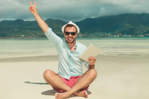 Człowiek siedzi na plaży z ipad w ręku Wyświetlono znak zwycięstwo — Zdjęcie stockowe