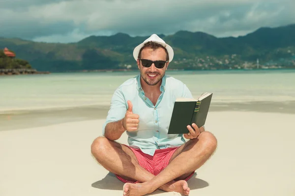 Homme assis sur la plage avec livre montrant signe de succès — Photo