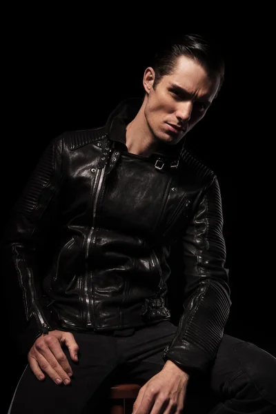Uomo in giacca di pelle nera in posa seduta in scuro studio backgro — Foto Stock