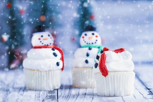 氷のクリスマススノーマンカップケーキの一部を噛んだり 食べています ニンジンの鼻 サンタの帽子 スカーフを持つ他の雪だるま 背景がぼやけた選択的フォーカス — ストック写真