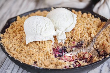 Böğürtlenli ve yaban mersinli tart dökme demir bir tavada pişmiş ve üzerinde vanilyalı dondurma ve kırsal beyaz ahşap bir masa. Bulanık arkaplan ile aşırı seçici odak.