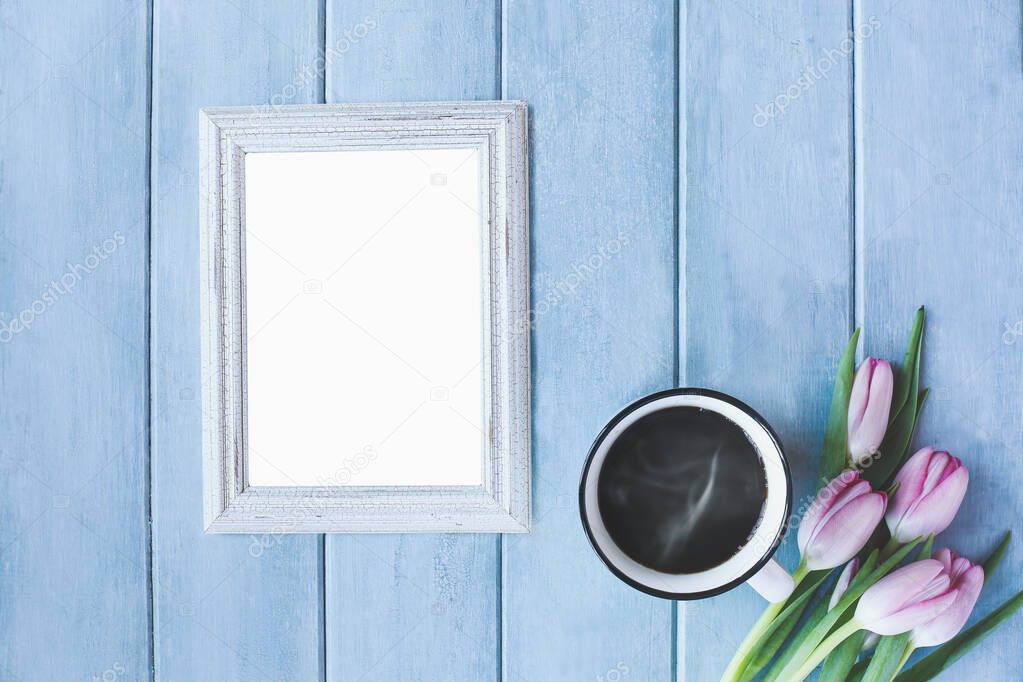 Vertical empty picture frame with steaming hot cup of black coffee and pink spring tulip flowers over a rustic blue wooden table shot from top view.
