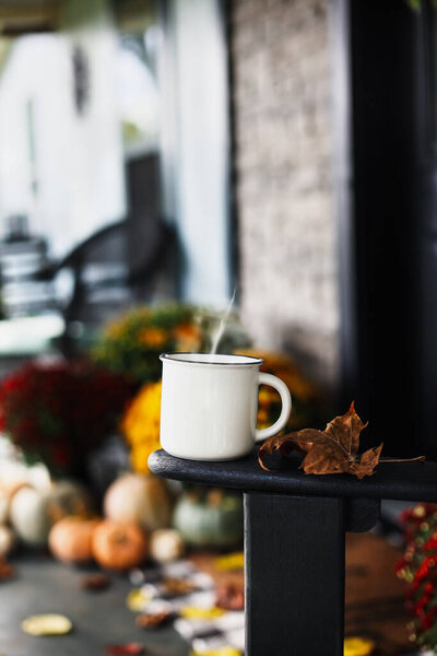 Hot steaming coffee sitting on rocking chair on front porch that has been decorated for autumn with heirloom white, orange, grey pumpkins, rain boots and mums. Selective focus with blurred background.