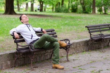 Young man sitting on the bench in the park and enjoying beautifu clipart