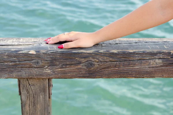 Female hand with red nails on wooden railing against green sea water in summer — Stock Photo, Image