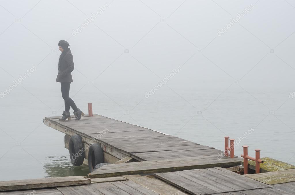 Lonely sad girl standing on wooden dock and watching river flow ??? Stock