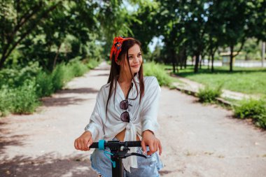 Parkta elektrikli scooter 'a binen tatlı esmer kız beyaz bir gömlek giyiyor. eko taşıma ve kira kavramı. 