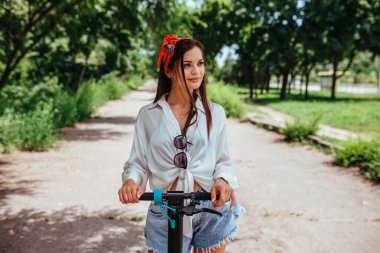Parkta elektrikli scooter 'a binen tatlı esmer kız beyaz bir gömlek giyiyor. eko taşıma ve kira kavramı. 