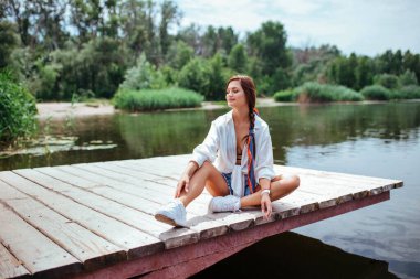 Beyaz gömlekli ve kot şortlu güzel bir esmer tahta bir köprüde oturuyor. Seyahat ve yürüme kavramı. 