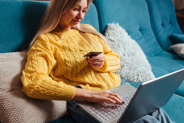 黄色のセーターを着た女性がソファに座ってクレジットカードを運転しています 家に座って買うという概念は — ストック写真