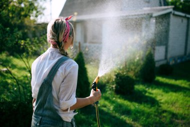a young woman in a white shirt and denim overalls watering the lawn. gardening concept and landscape design