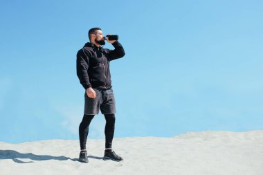 Şişirilmiş sporcu su içer ve kumlu bir dağda koştuktan sonra dinlenir.