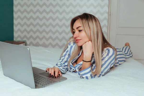 Une belle fille en pyjama est allongée sur le lit derrière un ordinateur portable. le concept du télétravail à domicile et du shopping — Photo