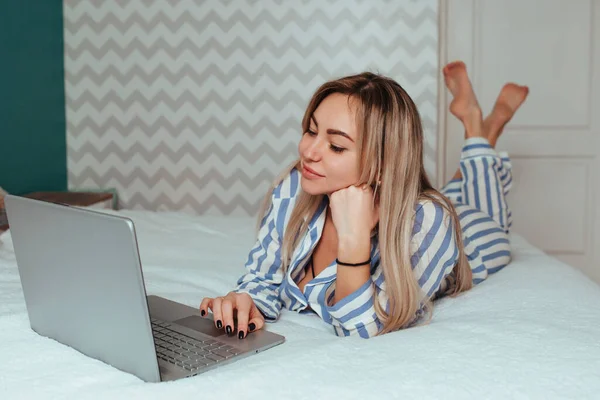 Een mooi meisje in pyjama ligt op het bed achter een laptop. het concept van thuiswerken op afstand en winkelen — Stockfoto