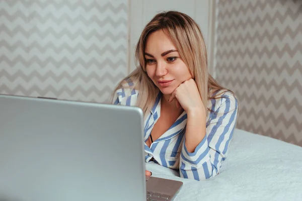 Een mooi meisje in pyjama ligt op het bed achter een laptop. het concept van thuiswerken op afstand en winkelen — Stockfoto