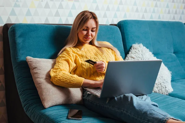Une femme avec un ordinateur portable dans un pull jaune s'assoit sur le canapé et conduit une carte de crédit donnée. le concept d'achat assis à la maison — Photo