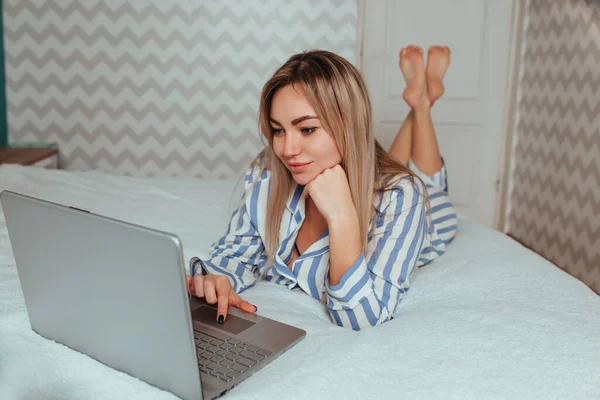 Een mooi meisje in pyjama ligt op het bed achter een laptop. het concept van thuiswerken op afstand en winkelen — Stockfoto