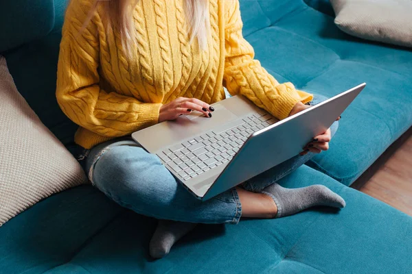 Een vrouw met een laptop in een gele trui zit op de bank. het concept van werken op afstand en winkelen terwijl u thuis zit — Stockfoto
