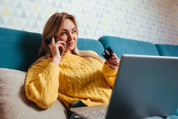 Een vrouw met een laptop in een gele trui zit op de bank en rijdt met een bepaalde creditcard. het concept van kopen terwijl je thuis zit — Stockfoto