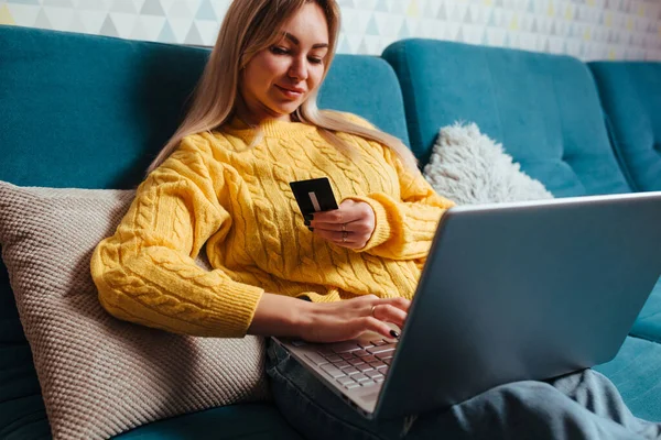 Een vrouw met een laptop in een gele trui zit op de bank en rijdt met een bepaalde creditcard. het concept van kopen terwijl je thuis zit — Stockfoto