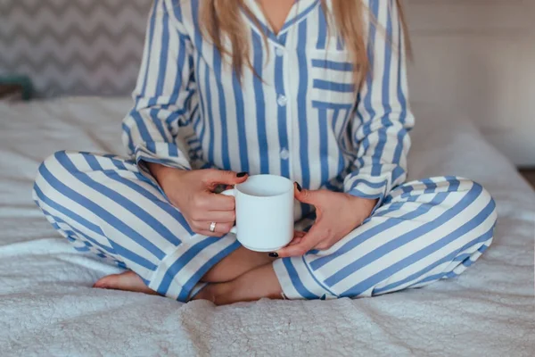 På morgonen sitter en tjej i pyjamas på sängen med en kopp kaffe i händerna. — Stockfoto